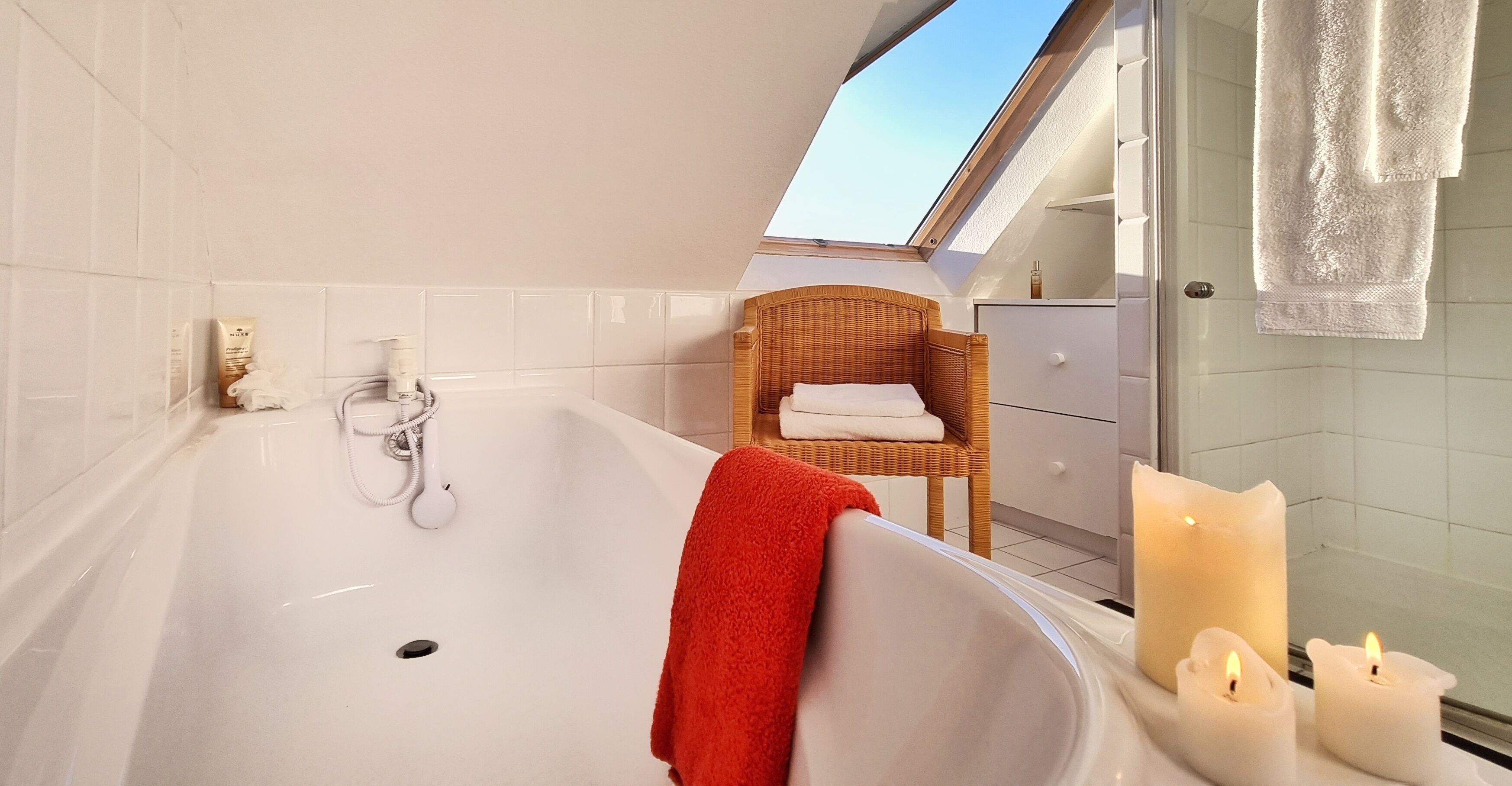 A modern bathroom with a large bathtub, a wicker chair, a glass shower enclosure, and a skylight.