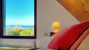 A bedroom with a large window overlooking a coastal landscape, a bedside lamp, and a red pillow.