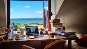 A desk with a laptop, books, and a glass of water near a window overlooking a beach.
