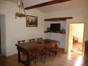 A dining area with a wooden table and chairs, a hanging light fixture, a painting on the wall, and a television on a shelf. - A gite of character and grand comfort in the heart of nature (Saint-Laurent-Le-Minier)