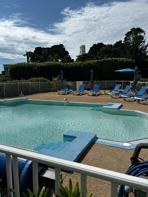 A swimming pool with lounge chairs and umbrellas, surrounded by a fence and greenery.