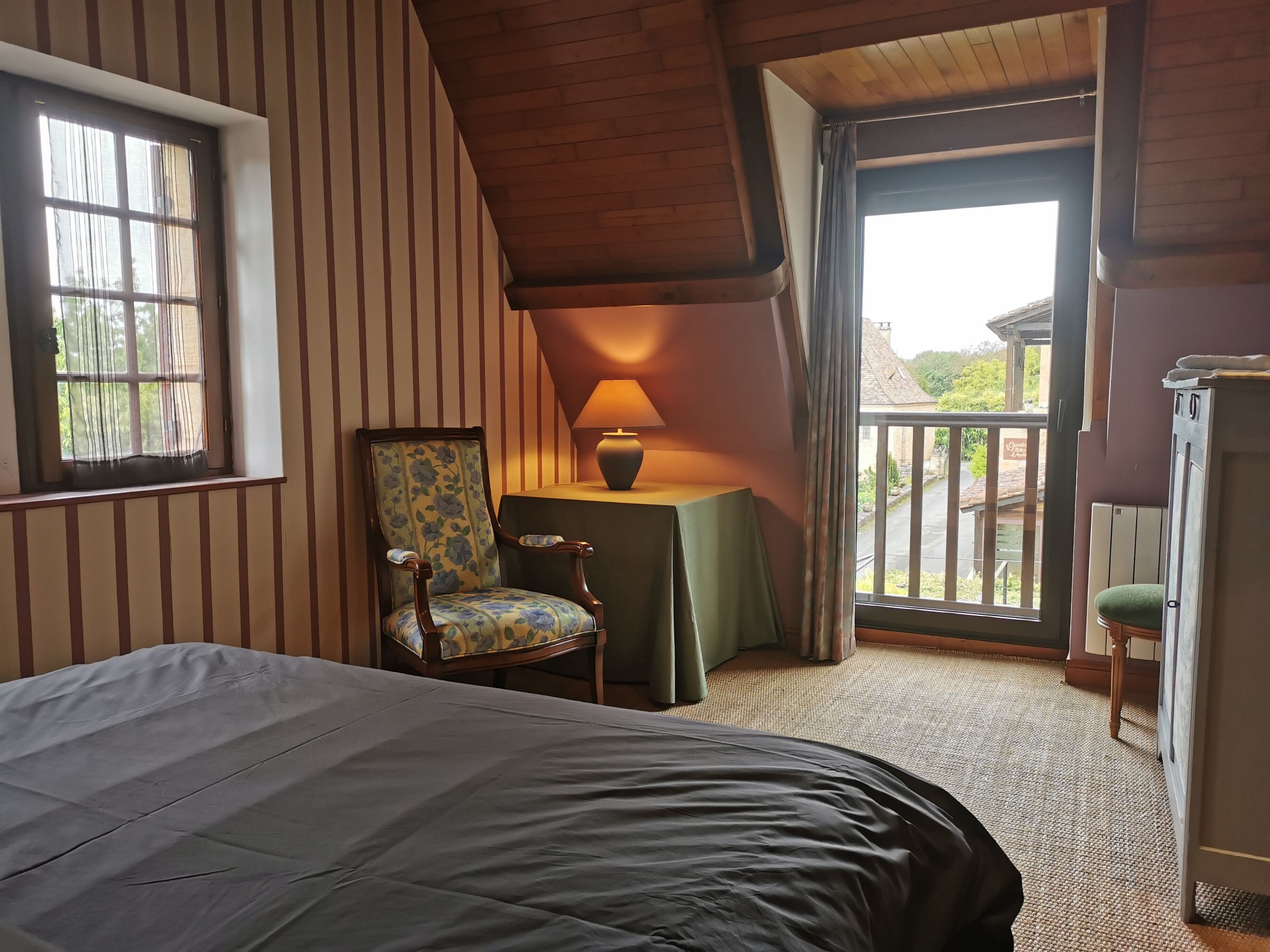 A bedroom with a bed, a chair, a table with a lamp, and a view of the outside.