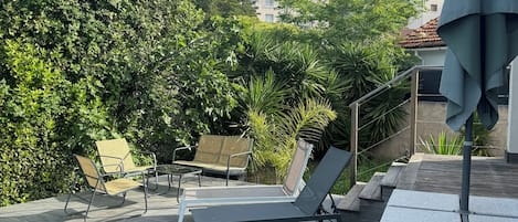 Et udendørs opholdsområde med loungesenge og et bord, omgivet af grønt og en swimmingpool.