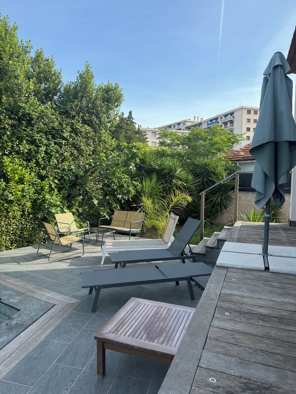 An outdoor seating area with lounge chairs and a table, surrounded by greenery and a pool.