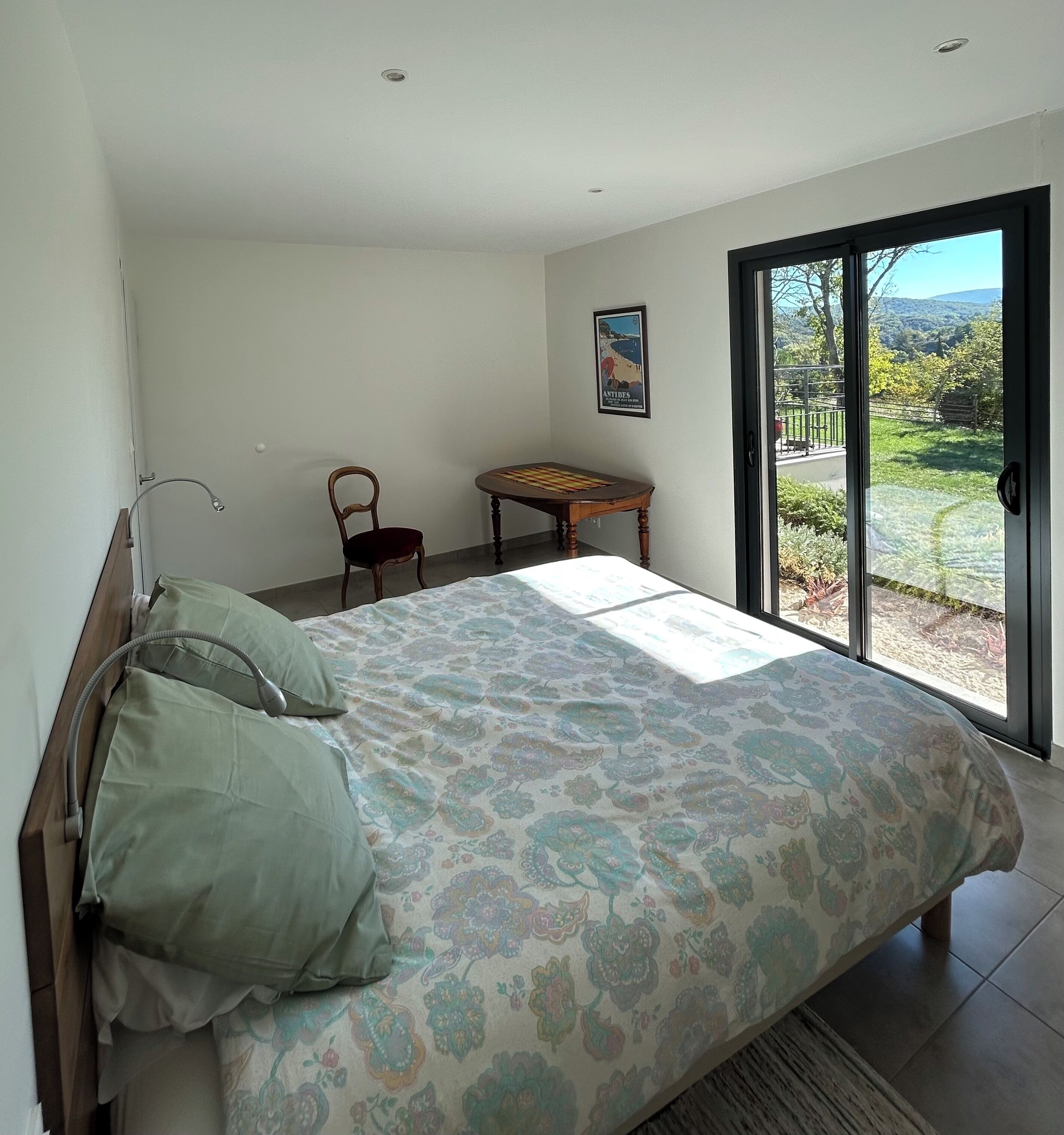 Een slaapkamer met een bed, een stoel, een kleine tafel en een raam met uitzicht op groen.