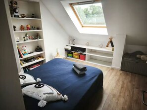 Una camera da letto con soffitto spiovente, una finestra a lucernario, un letto con copriletto blu, una mensola con giocattoli e una scrivania con una lampada.