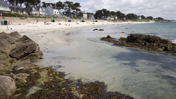 A sandy beach with clear water, rocky outcrops, and houses in the background.