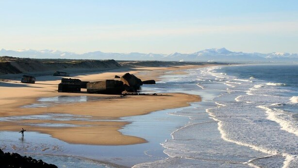 Een strand met grote rotsen, een zandstrand en bergtoppen in de verte.