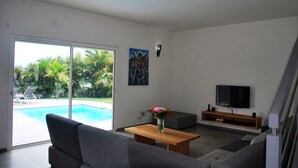 Un salon moderne avec vue sur la piscine, doté de canapés gris, d’une table basse en bois et d’un téléviseur à écran plat.