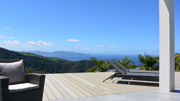 Une terrasse en bois avec une chaise en osier et un fauteuil de relaxation, offrant une vue sur un paysage montagneux et la mer.