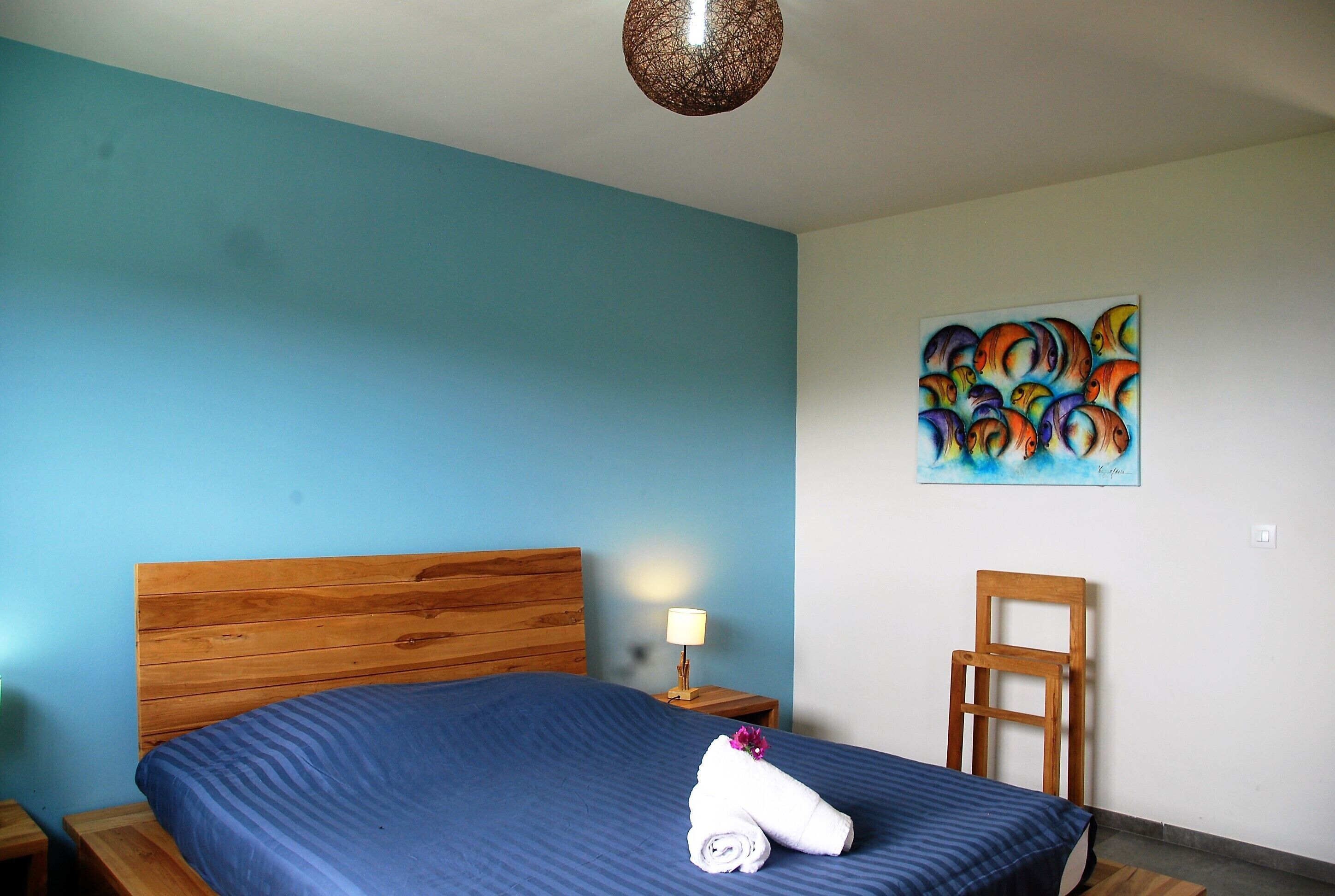 A neatly made bed with a blue bedspread, a wooden headboard, a wooden nightstand with a lamp, a wooden chair, and a colorful abstract painting on the wall.