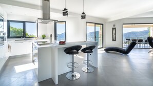 A modern kitchen with a central island, bar stools, and large windows offering a view of the mountains. - Spacious villa, oustanding views on sea and mountains, garden is 1600m2, 5**** (Les Adrets-De-L'estérel)