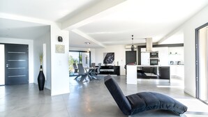 A spacious living area with a modern kitchen, a dining table, and a black lounge chair. - Spacious villa, oustanding views on sea and mountains, garden is 1600m2, 5**** (Les Adrets-De-L'estérel)