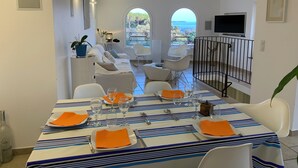 A dining area with a table set for four, white chairs, and a view of the sea through arched windows.