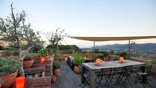 A wooden deck with a table and chairs, surrounded by potted plants and a barbecue grill, overlooking a scenic landscape.