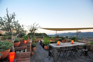 Une terrasse en bois avec une table et des chaises, entourée de plantes en pot et d’un barbecue, offrant une vue sur un paysage pittoresque.