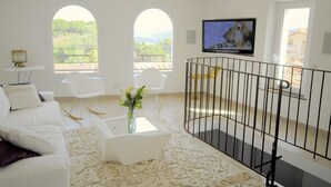 A modern living room with a white sofa, a coffee table with a vase, and a TV on the wall. There are two arched windows offering a view of a balcony with a railing.