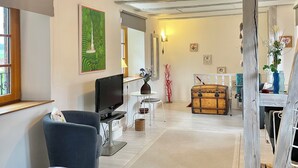 Een woonkamer met een televisie, een bank, een eethoek met een tafel en stoelen, een houten kist, en een trap naar een ander niveau.