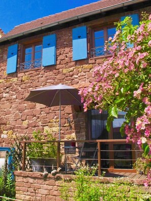A stone building with blue shutters, a patio with a table and chairs, and a pink flowering tree.