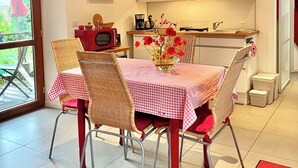 Een eetgedeelte met een geruit tafelkleed, rieten stoelen en een vaas rode bloemen.
