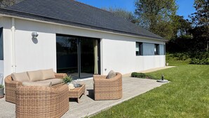 Una casa moderna con un amplio ventanal corredero, un patio con mobiliario de mimbre y un césped bien cuidado.
