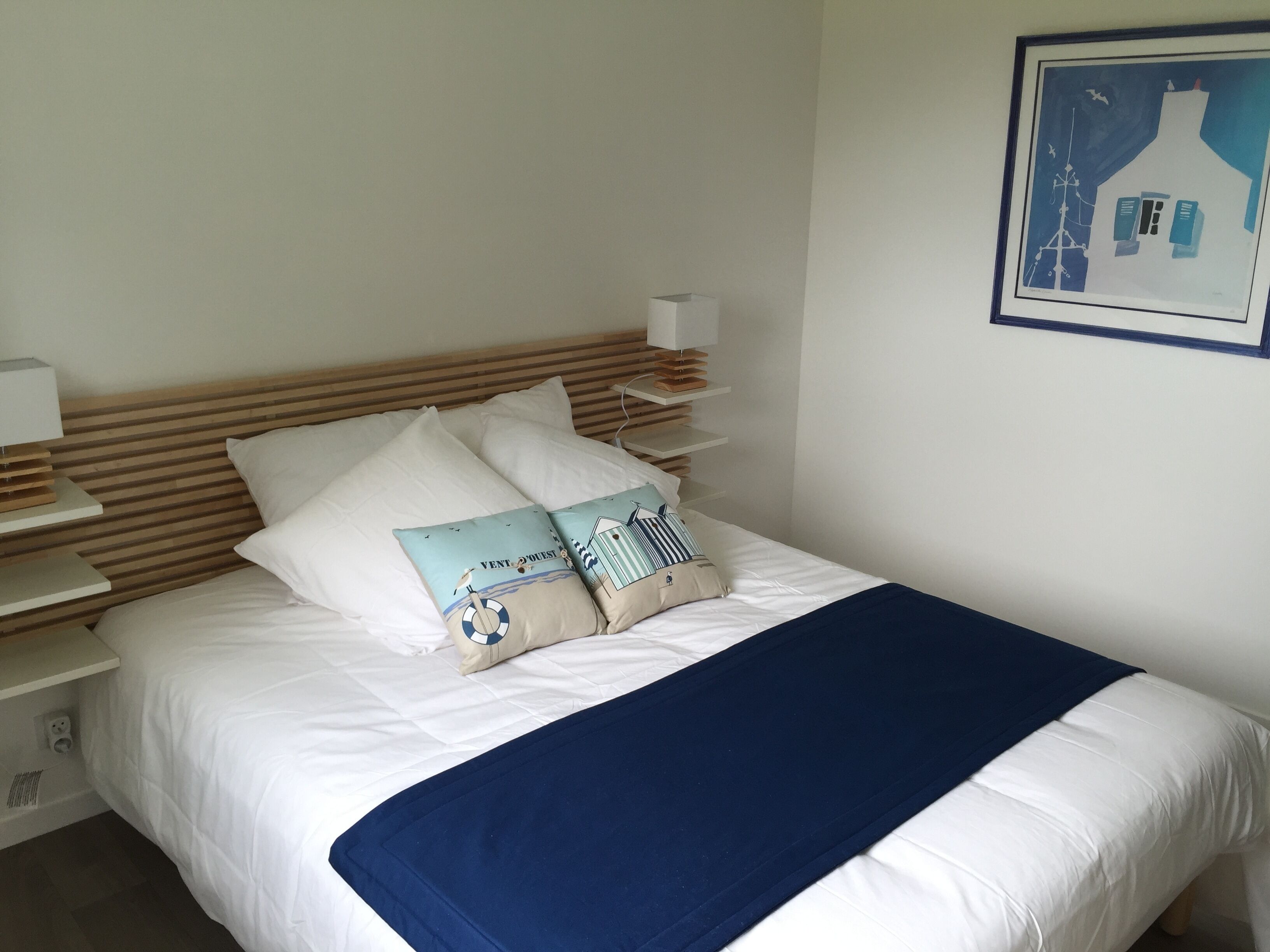 Ein ordentlich bezogenes Bett mit weißer Bettwäsche und einer blauen Tagesdecke. Zwei dekorative Kissen mit maritimen Motiven. Ein gerahmtes Bild hängt an der Wand über dem Bett.