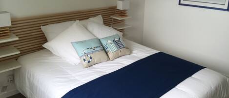 Ein ordentlich bezogenes Bett mit weißer Bettwäsche und einer blauen Tagesdecke. Zwei dekorative Kissen mit maritimen Motiven. Ein gerahmtes Bild hängt an der Wand über dem Bett.