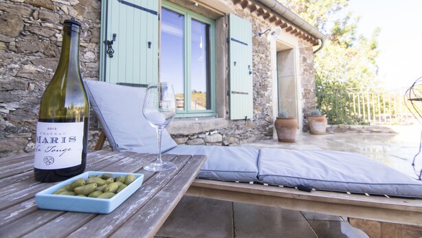 A bottle of wine, a glass, and a bowl of olives on a wooden table outside a stone building with green shutters.