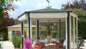 A covered outdoor seating area with a glass table and chairs, surrounded by potted plants and a pool in the background. - Country cottage fully furnished, in private property, quiet. (Draguignan)