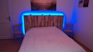 Un dormitorio con cabecera de madera, dos sillas de madera, una cama con edredón blanco y un cuadro en la pared.