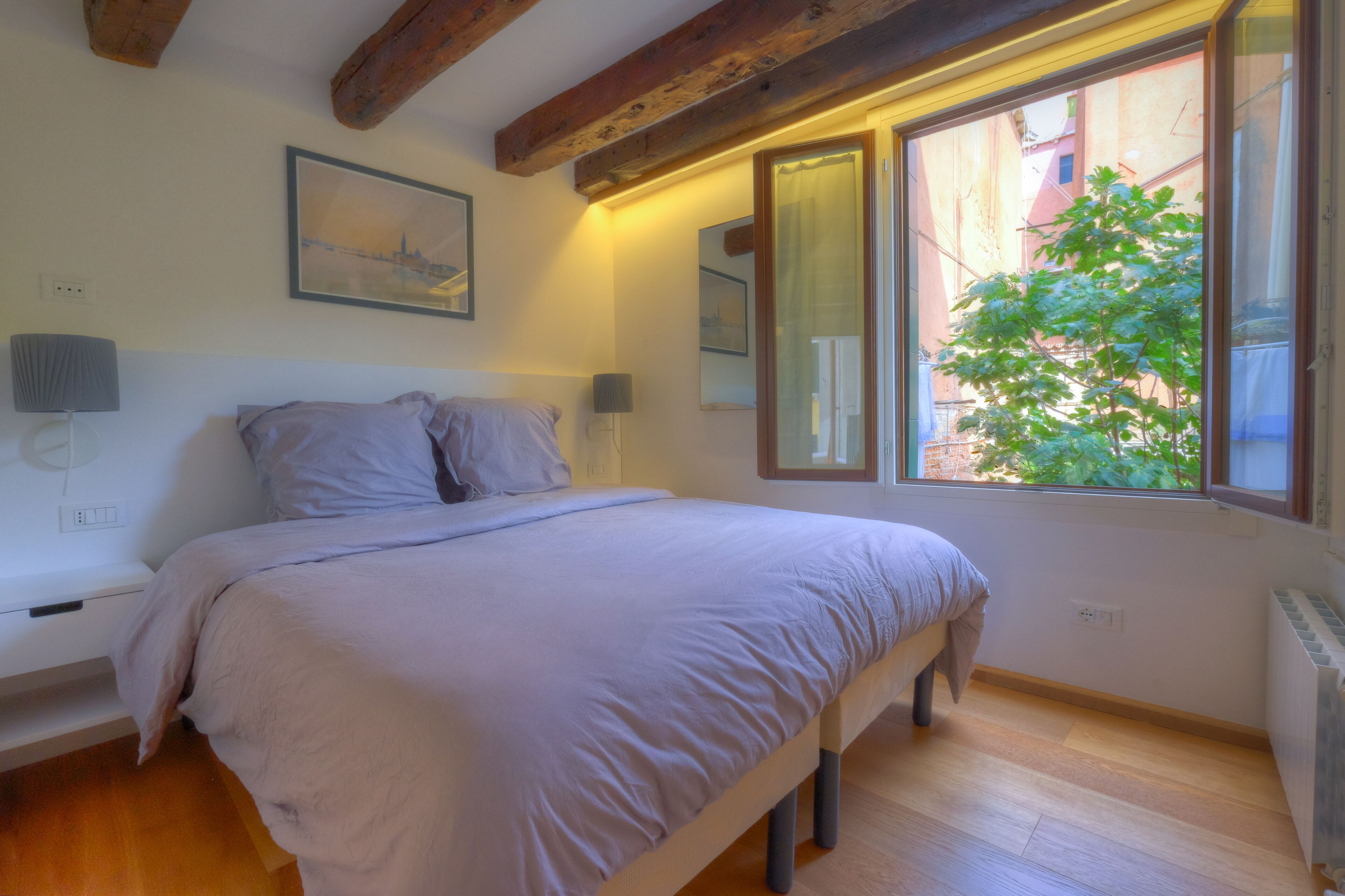 Un dormitorio con cama, lámparas de noche, una mesita de noche, una ventana con vista a edificios y un cuadro en la pared.