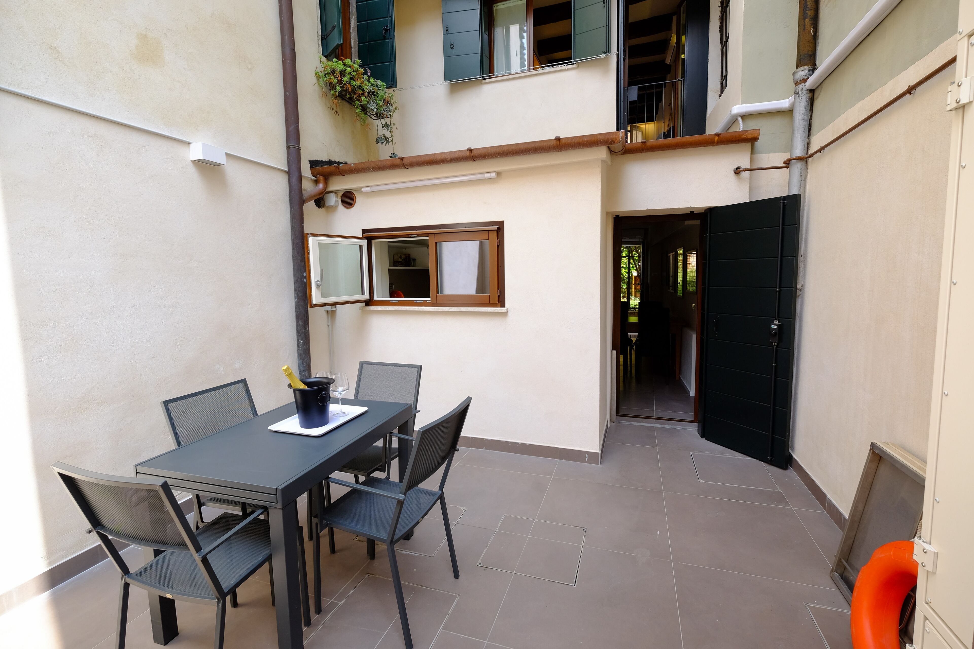 Un pequeño espacio exterior para comer con mesa y sillas, una ventana y una puerta que da a un corredor cubierto.