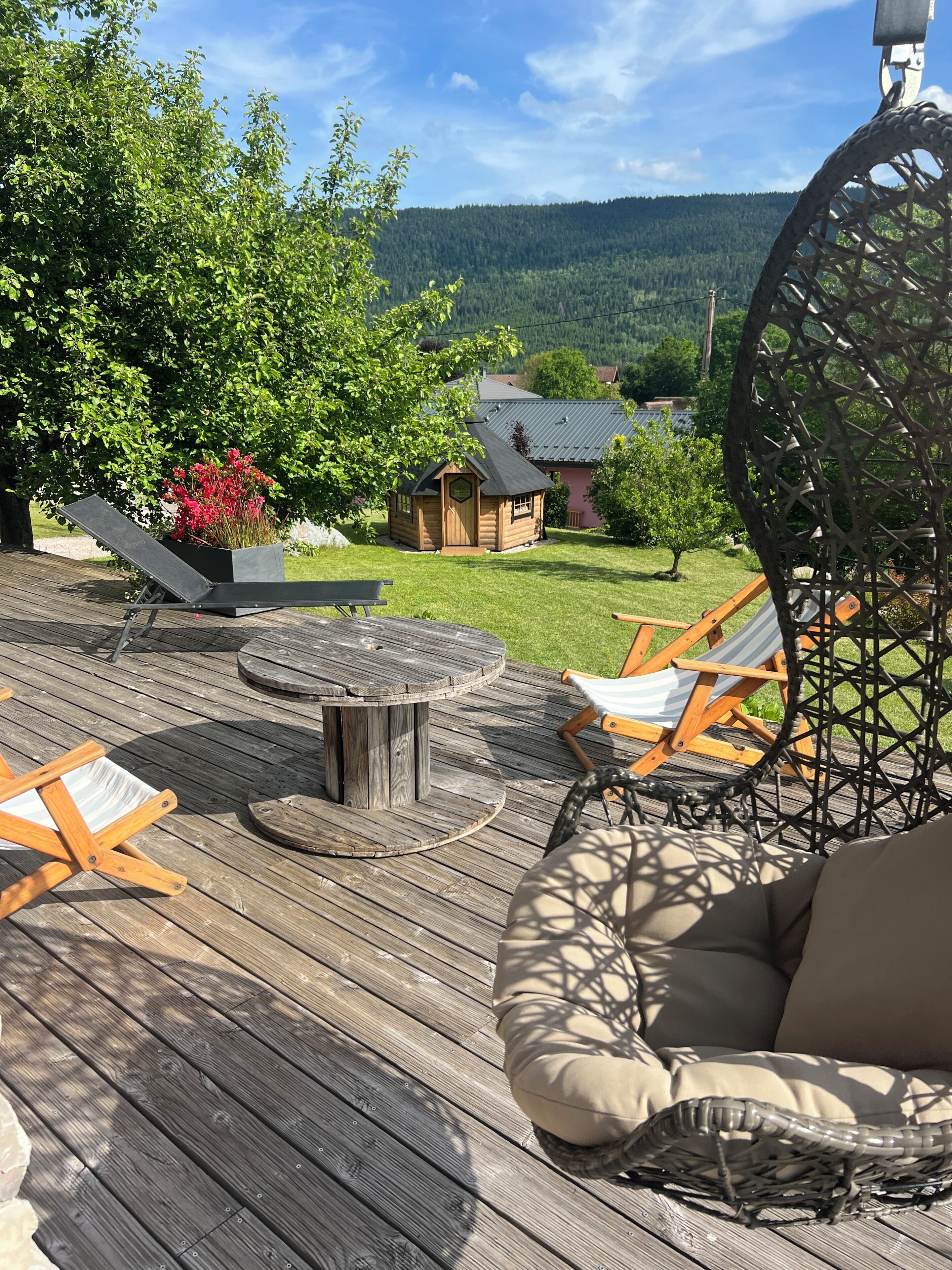 Een houten terras met een ronde tafel, twee loungestoelen, een hangstoel en een klein gebouw met een schuin dak.