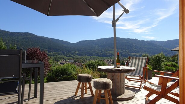 Une terrasse en bois avec des meubles d’extérieur, une table avec une bouteille et des verres, et une vue sur des montagnes et une ville en arrière-plan.