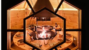 Une pièce en bois de forme hexagonale, dotée d’un foyer central, entourée de tables et de chaises en bois.
