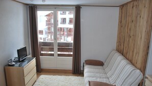 Una habitación pequeña y luminosa con un sofá, un televisor sobre un mueble de madera y vistas a edificios por una puerta corrediza.