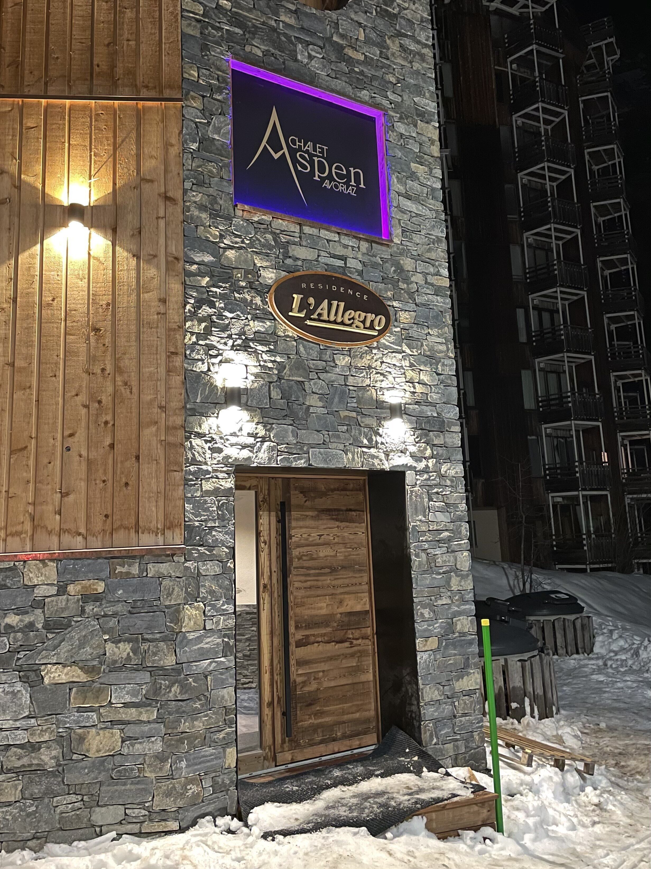 Ein Gebäude mit einer Steinaußenwand, einer Holztür und Schildern mit den Aufschriften 'Chalet Aspen' und 'L'Allegro Residence'.