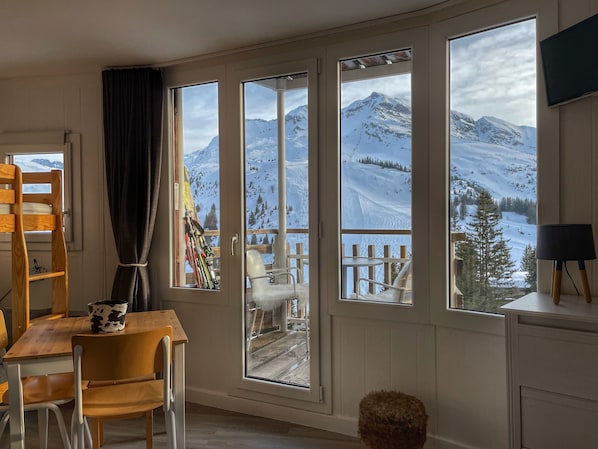 Une pièce avec une table et des chaises en bois, un porte-skis et de grandes fenêtres donnant sur des montagnes enneigées.