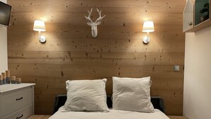 A hotel room with a wooden headboard, two bedside lamps, a bed with white linens, a small bedside table, and a dresser with drawers. - Charming Studio 4 pers with south balcony and mountain view (Morzine)