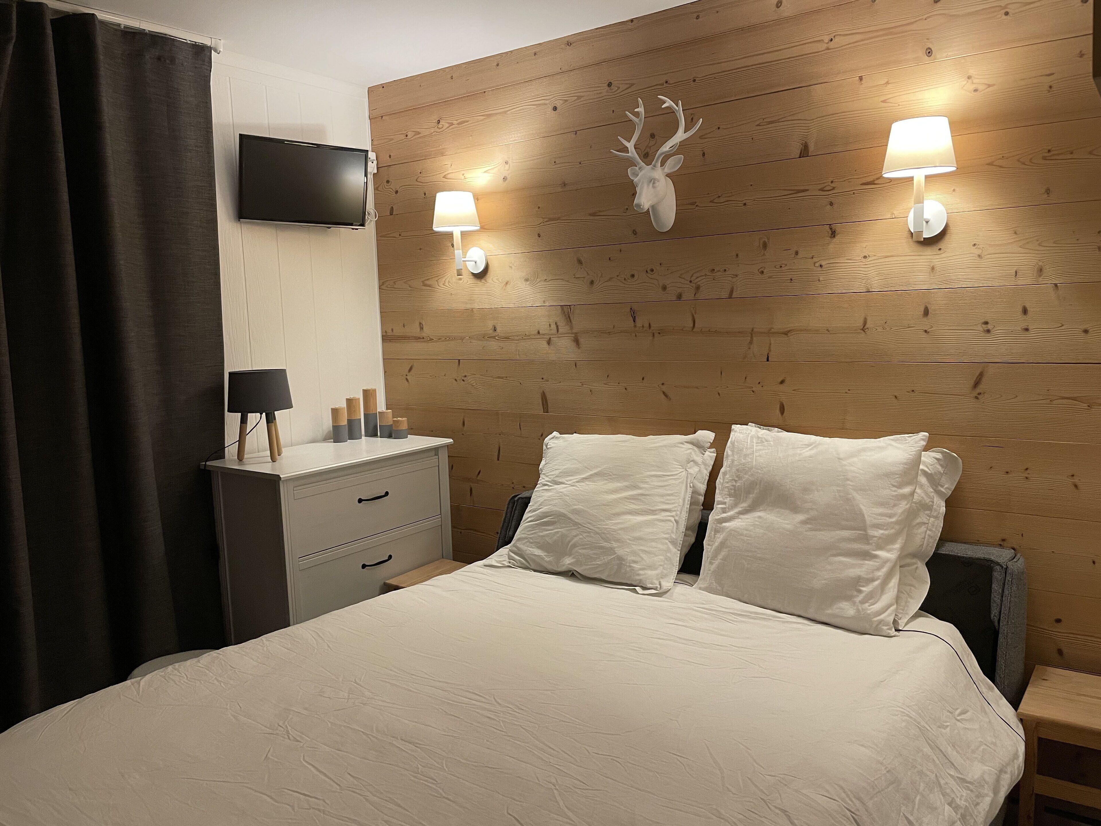Un dormitorio con cabecera de madera, dos lámparas de noche, un televisor montado, una mesita de noche con lámpara y una cama con ropa de cama blanca.
