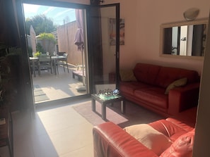 An open sliding glass door leading to an outdoor patio with a dining table and chairs, a red leather sofa inside, and a mirror on the wall. - BEAUTIFUL air-conditioned T2 60m2 100 m from Les Lecques beach + parking lot (Saint-Cyr-Sur-Mer)