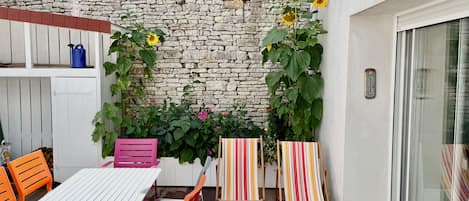 Un patio al aire libre con una mesa blanca y sillas naranjas, un muro de piedra y plantas en macetas.