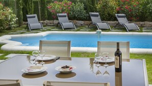 Zona de comedor junto a la piscina con una mesa para dos, sillas y una botella de vino.