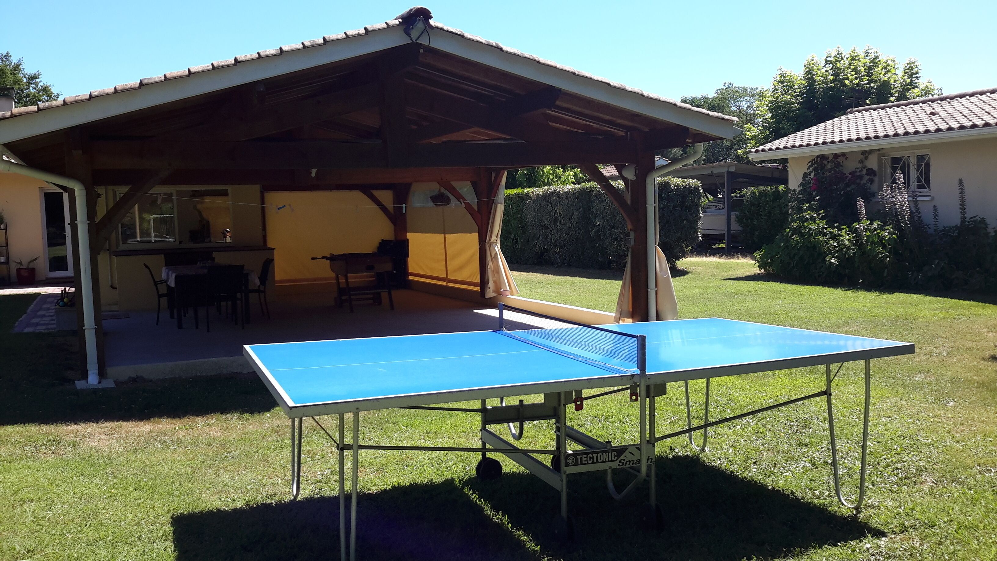 Una mesa de ping pong al aire libre bajo un pabellón de madera, con una mesa y sillas cerca.