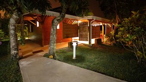 A well-lit outdoor area with a pathway leading to a wooden structure, surrounded by lush greenery and trees.