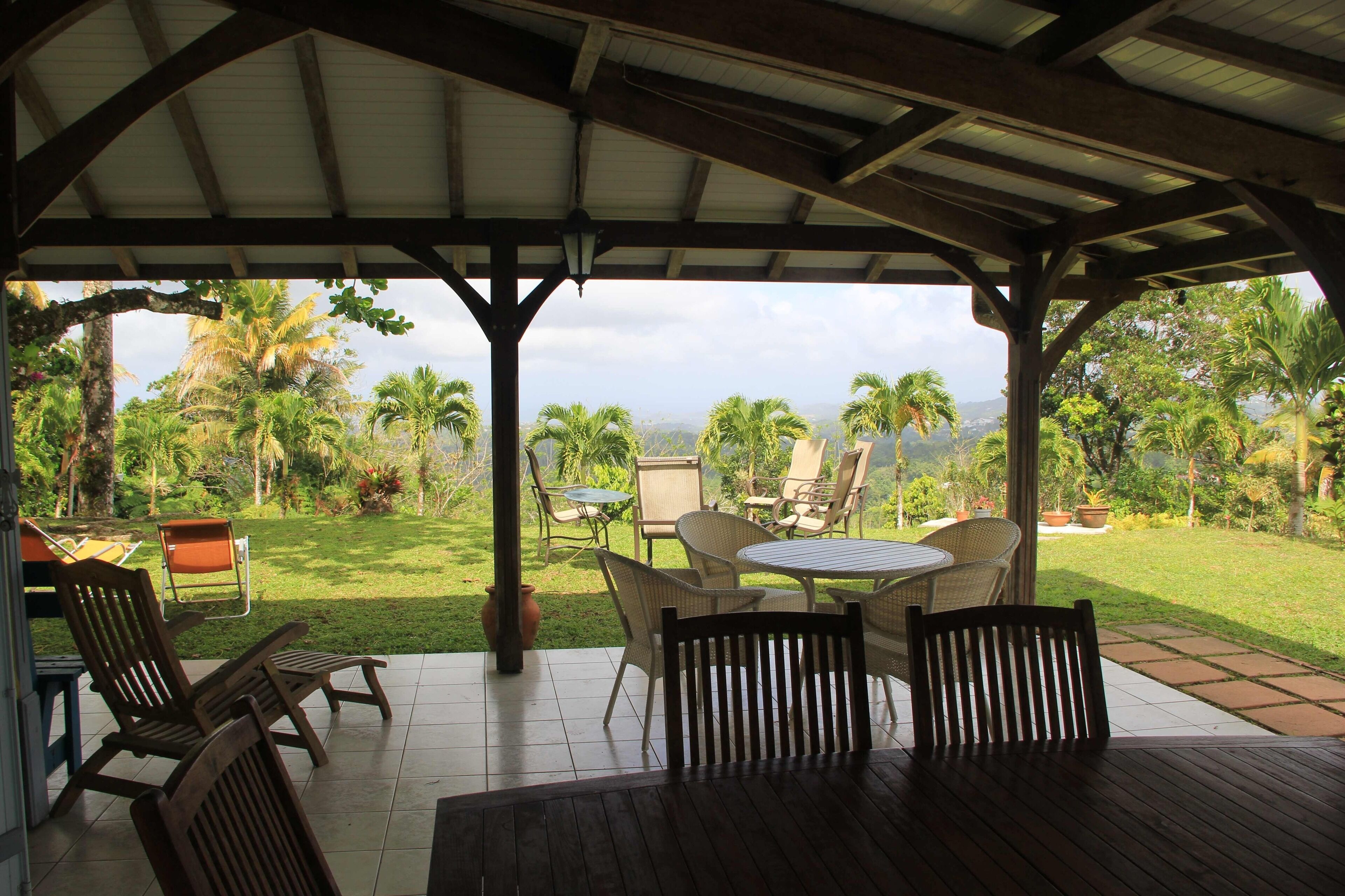 An outdoor patio with wooden furniture, a covered area, and a view of a lush garden with palm trees.