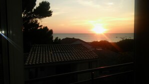 Vista al atardecer desde un balcón con vistas a la costa, techos de tejas y árboles.