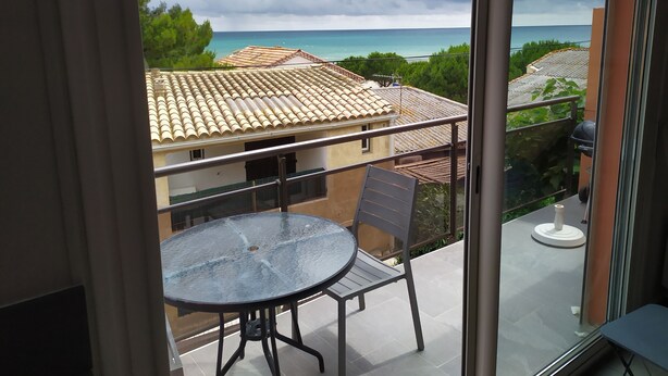 Balcón con mesa y sillas de vidrio, con vista a una zona costera con edificios y al mar.
