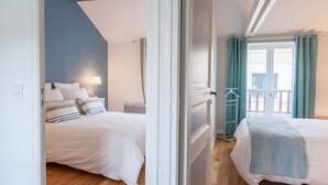 A bedroom with two beds, a wardrobe, a window with curtains, and a view of buildings outside.