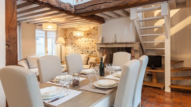 A dining area with a wooden table set for a meal, surrounded by chairs, with a stone wall and a staircase in the background.